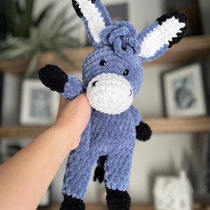 May include: A handmade, plush donkey toy in shades of blue, white, and black. The donkey has large ears with white and black accents, a white muzzle, and black hooves. The toy is made of soft, textured yarn and is designed to be cuddly.