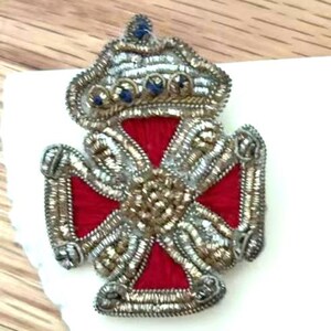 May include: A crown-topped cross patch featuring vibrant red fabric embroidery accented with metallic silver and gold bullion wire thread. The crown is embellished with small blue beads, and the entire piece is outlined in intricate silver wire work.