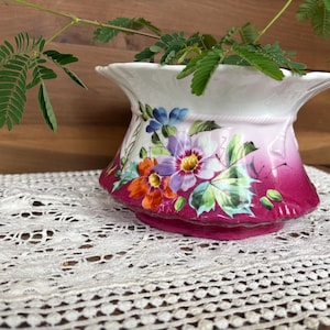 May include: A white ceramic planter with a flared rim and a deep magenta base featuring a painted floral design of purple, orange, and blue flowers with green leaves. A small green plant with delicate, feathery leaves grows from the center.