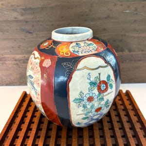 Könnte beinhalten: Eine runde Keramikvase mit glänzender Oberfläche und abwechselnden Feldern in Tiefschwarz und leuchtendem Rot. Die Felder sind mit detaillierten, handgemalten Blumenmotiven in Blau-, Grün- und Orangetönen verziert, akzentuiert durch Goldlinien.