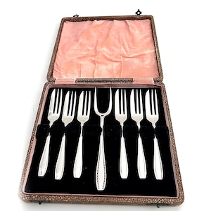 Könnte beinhalten: Ein Set Silberbesteck in einem Vintage-Etui. Die Gabeln sind in einer mit schwarzem Samt ausgekleideten Box mit rosa Satin-Innenfutter angeordnet. Das Etui ist braun mit einem strukturierten Muster und einem Verschluss.