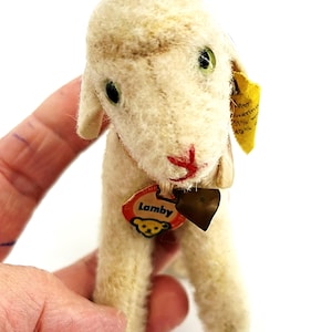 Könnte beinhalten: Ein Vintage-Lamm-Stofftier mit cremefarbenem Körper, grünen Augen und einem rot genähten Mund. Es hat eine Glocke um den Hals und ein Etikett mit der Aufschrift "Lamby". Das Lamm wird in einer Hand gehalten.