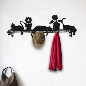 Könnte beinhalten: Schwarzes Metall-Wandregal mit Katzen- und Blumensilhouetten. Ein Strohhut und eine rote Jacke hängen darunter. Eine braune Tragetasche steht auf einer Holzbank.