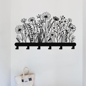 Op de afbeelding: Zwart-witte wanddecoratie met een bloemenmotief en daaronder een zwart metalen haakrek. Het haakrek heeft vijf haken.