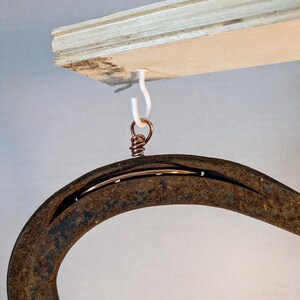 May include: A rusty horseshoe hanging from a white hook attached to a wooden board. The horseshoe is attached to the hook with a copper wire.