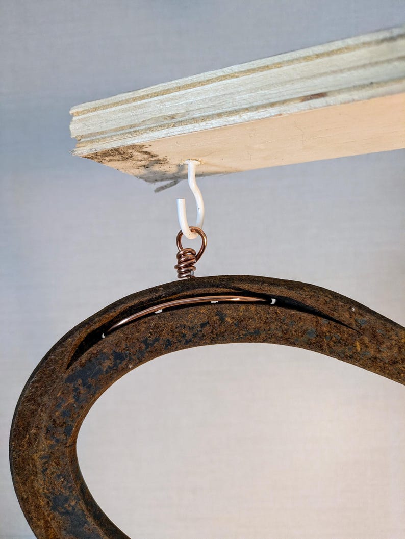 May include: A rusty horseshoe hanging from a white hook attached to a wooden board. The horseshoe is attached to the hook with a copper wire.
