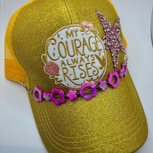 May include: Gold glitter baseball cap with a mesh back. The cap features a decorative pink star and a floral beaded chain. The front of the cap has the text "My Courage Always Rises" in a circular design.