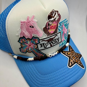 May include: A light blue and white trucker hat featuring a variety of embroidered patches including a pink horse, a cowgirl, a cross, a star with a leopard print, and a rectangular patch with the text Howdy, accented by a beaded hat band.