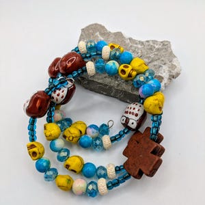 May include: A multi-strand bracelet featuring blue, yellow and brown beads. The beads are in various shapes, including skulls, owls and hearts. There is a brown cross charm on the bracelet.