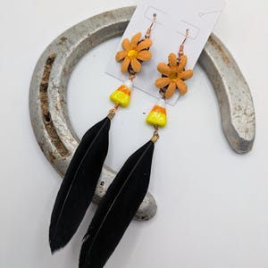 May include: A pair of dangle earrings with black feathers, orange and yellow candy corn charms, and orange flower charms.