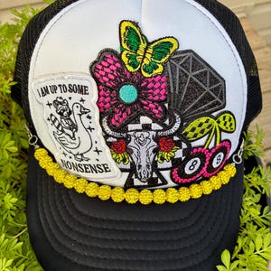 May include: A black and white trucker hat featuring a yellow beaded chain across the brim. The front panel displays embroidered patches including a raccoon riding a goose with the text I AM UP TO SOME NONSENSE, a butterfly, a skull, and cherries.