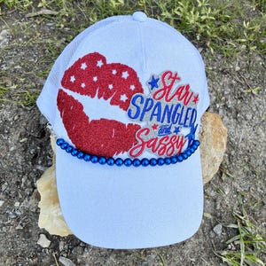 May include: White trucker hat with a mesh back and a curved brim. The front features a red, white, and blue design of lips and the text "Star Spangled and Sassy". A blue beaded chain accents the brim.