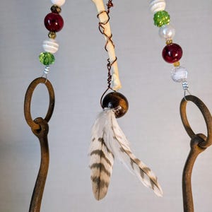 May include: A rustic metal horseshoe wind chime with beads, feathers, and a bone hanging from it. The beads are white, green, and red. The feathers are brown and white. The bone is white.