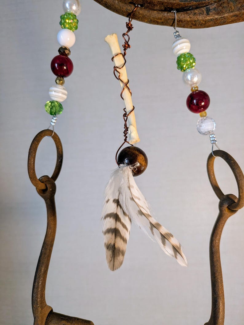 May include: A rustic metal horseshoe wind chime with beads, feathers, and a bone hanging from it. The beads are white, green, and red. The feathers are brown and white. The bone is white.