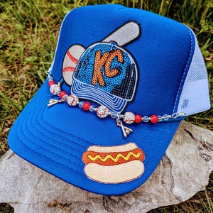 May include: A blue baseball cap with a white mesh back, featuring a baseball, a baseball bat, and a patch with "KC" on it. A beaded chain with baseball charms and a hot dog patch add detail.
