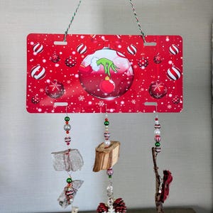 May include: A red and white Christmas ornament with a Grinch design hangs from a string of beads. The ornament is surrounded by smaller red and white ornaments and snowflakes. The ornament is attached to a red license plate with a white border.