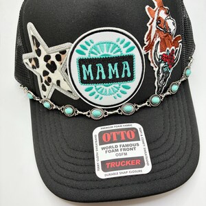 May include: Black trucker hat with a mesh back, featuring a star with a leopard print, a round patch with the word "MAMA", and a horse head with a skull. A turquoise beaded chain accents the brim. The hat has the text "OTTO TRUCKER".