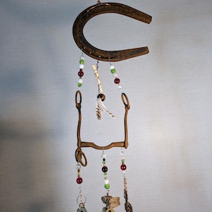 May include: A rustic wind chime made from a horseshoe, a bit, and keys. The chime features red, green, and white beads, as well as a feather.