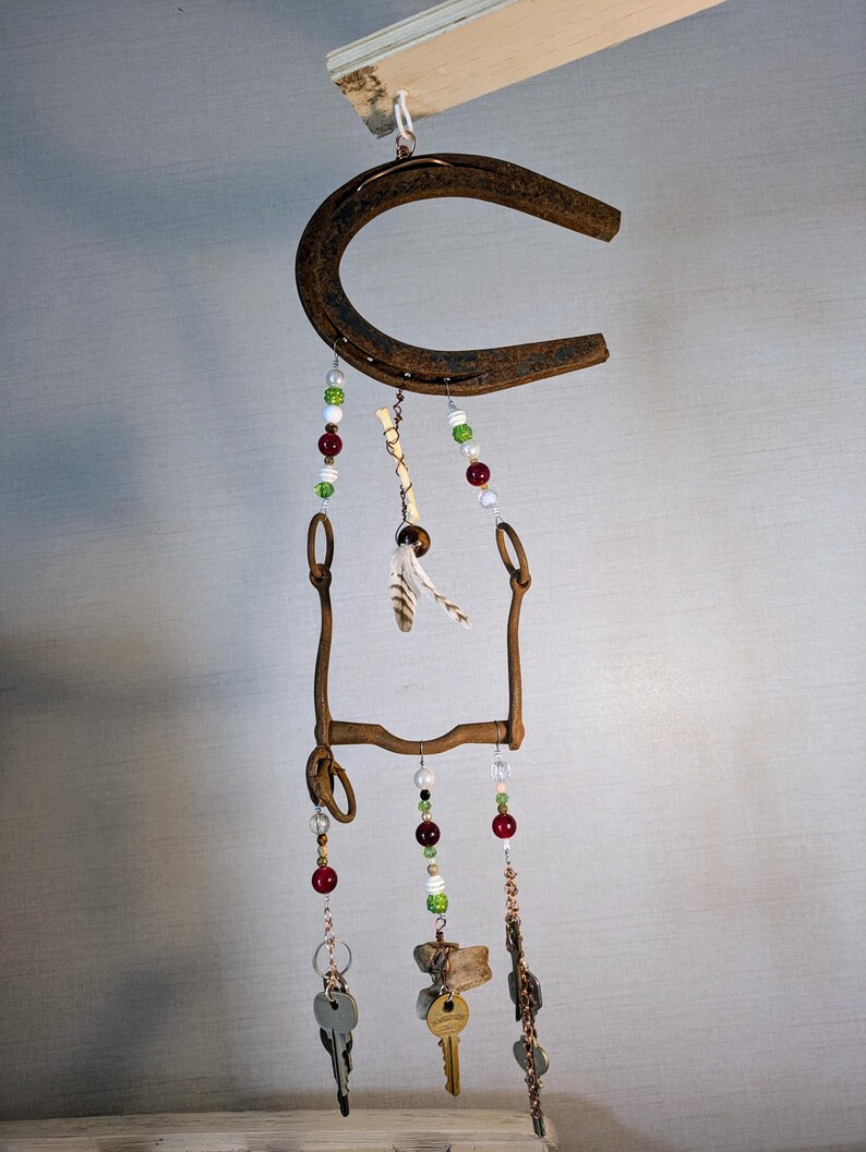 May include: A rustic wind chime made from a horseshoe, a bit, and keys. The chime features red, green, and white beads, as well as a feather.