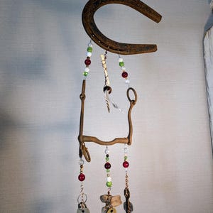 May include: A rustic horseshoe wind chime with beads and keys hanging from it. The horseshoe is rusty and has a brown patina. The beads are red, green, and white. The keys are silver and have a worn look. The wind chime is hanging from a wooden beam.