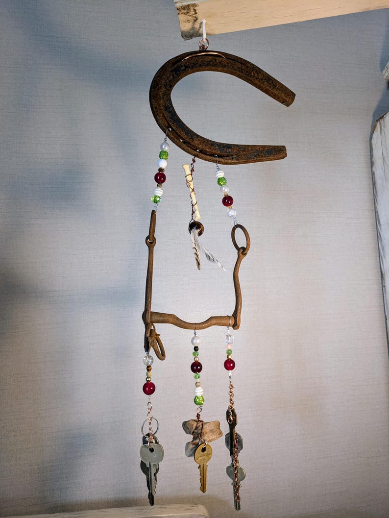 May include: A rustic horseshoe wind chime with beads and keys hanging from it. The horseshoe is rusty and has a brown patina. The beads are red, green, and white. The keys are silver and have a worn look. The wind chime is hanging from a wooden beam.