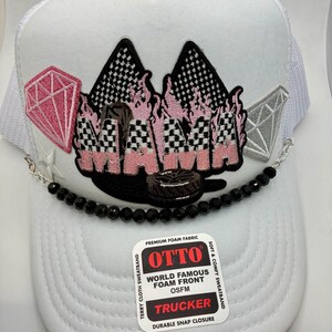 May include: White trucker hat with a mesh back and a foam front. The front features a pink and black embroidered design with a diamond and flame motif. A black beaded chain and a tag that says "OTTO" are also visible.