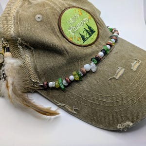 May include: A green and brown mesh baseball cap with a green embroidered patch that says "Happy Camper" and a beaded trim. The cap has a feather attached to the side.