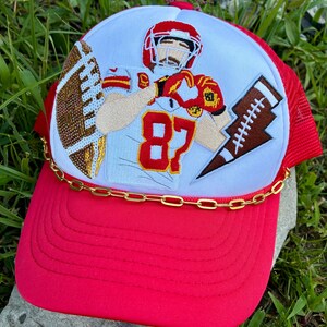 May include: Red and white trucker hat featuring an embroidered football player in a red and white uniform with the number 87. The hat has a red brim, white crown, and a gold chain detail.