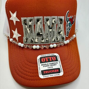 May include: Rust-colored trucker hat with a white mesh back. The word "MAMA" is embroidered in a cow print design. Features white star accents, a beaded chain, and a floral skull patch. The hat has a "TRUCKER" label.