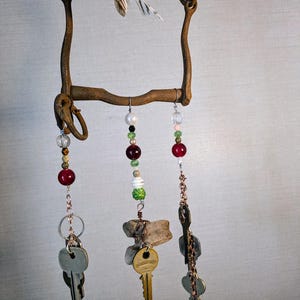 May include: A rustic metal wind chime with multiple strands of beads and keys hanging from it. The beads are red, green, white, and clear. The keys are silver and gold. The wind chime is made from a repurposed horse bit.