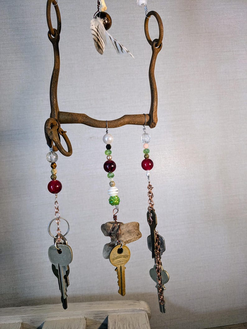 May include: A rustic metal wind chime with multiple strands of beads and keys hanging from it. The beads are red, green, white, and clear. The keys are silver and gold. The wind chime is made from a repurposed horse bit.