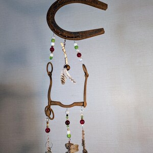 May include: A rustic horseshoe wind chime with beads, feathers, and keys hanging from it. The horseshoe is brown and rusty, and the beads are red, green, and white. The feathers are white and brown, and the keys are silver.