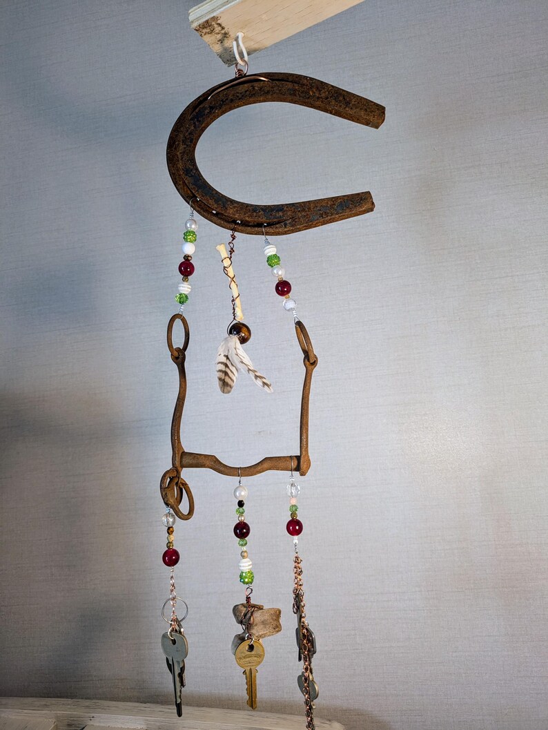 May include: A rustic horseshoe wind chime with beads, feathers, and keys hanging from it. The horseshoe is brown and rusty, and the beads are red, green, and white. The feathers are white and brown, and the keys are silver.