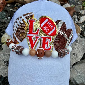 May include: White baseball cap with the word "LOVE" in red and gold, adorned with football-themed embellishments. Features a beaded band with football and white beads. The cap has a glittery texture.