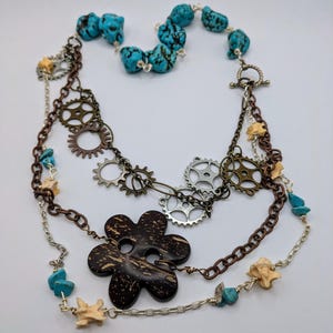 May include: A multi-strand necklace with turquoise stones, metal gears, and a brown wooden flower pendant.