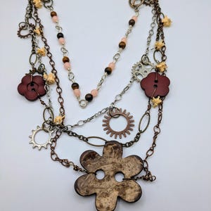 May include: A multi-layered necklace with brown, pink, and silver beads, metal chains, and various charms, including gears, flowers, and buttons. The necklace features a large brown wooden flower charm.