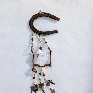 May include: A rusty horseshoe with a metal frame and beads hanging from it. The beads are red, green, and white. There are also feathers and keys hanging from the frame.