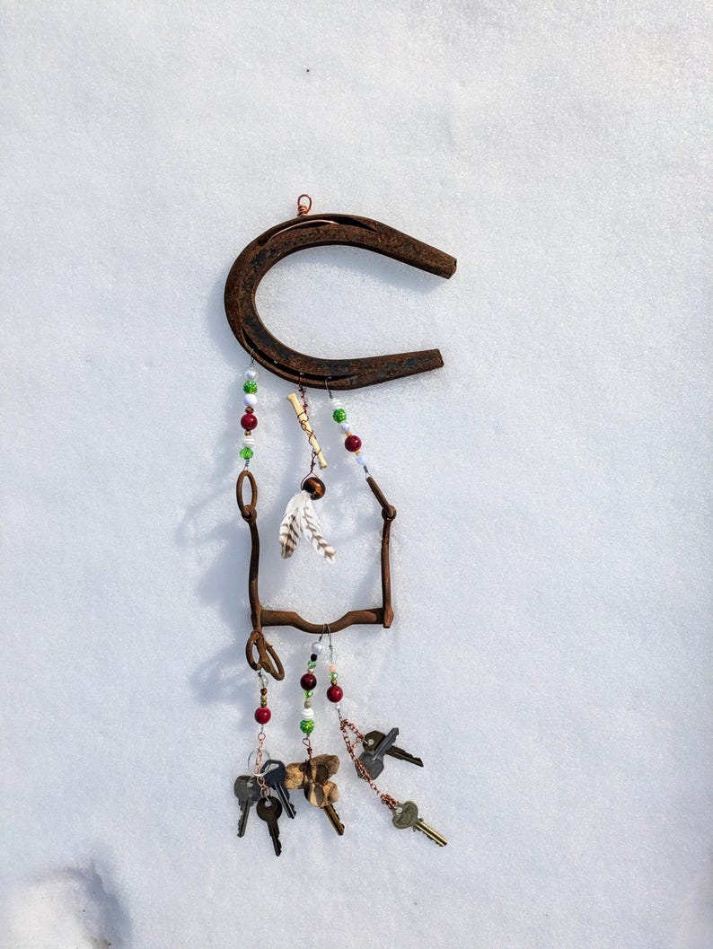 May include: A rusty horseshoe with a metal frame and beads hanging from it. The beads are red, green, and white. There are also feathers and keys hanging from the frame.