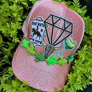 May include: A pink glitter trucker hat featuring a patch with a horse and the text "SAVE WATER drink marg", a diamond shape, a bull skull, a margarita glass, and a decorative chain of small green cactus beads across the front panel.
