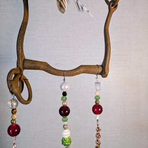 May include: A rustic metal horse bit with a feather charm and dangling beads in shades of red, green, white, and gold. The beads are strung on copper chains and wire.