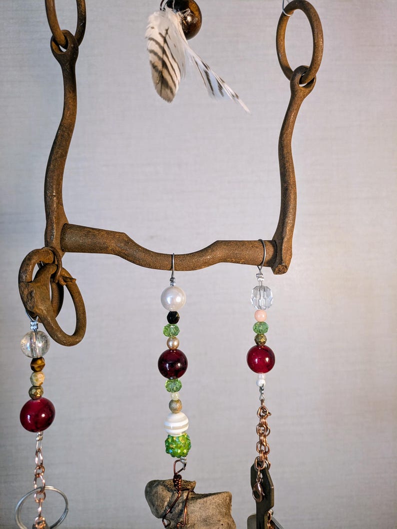 May include: A rustic metal horse bit with a feather charm and dangling beads in shades of red, green, white, and gold. The beads are strung on copper chains and wire.