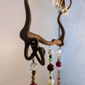 May include: A rustic metal wind chime with a horseshoe-shaped frame and multiple strands of beads. The beads are in various colors, including red, green, white, and brown. A single white feather hangs from the top of the frame.