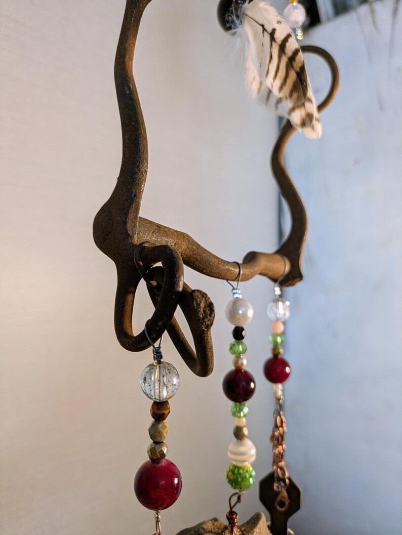 May include: A rustic metal wind chime with a horseshoe-shaped frame and multiple strands of beads. The beads are in various colors, including red, green, white, and brown. A single white feather hangs from the top of the frame.