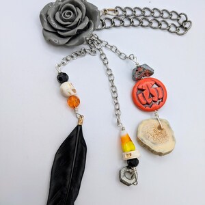 May include: A gray rose charm with a silver chain and various Halloween-themed charms, including a pumpkin, a candy corn, a skull, a feather, and a piece of antler.