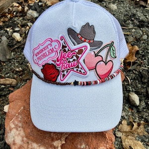 May include: A white trucker hat with a mesh back and a curved brim. The hat is decorated with patches including a cowboy hat, cherries, a star, and the text "Somebody's Problem" and "Yee Haw". A beaded band accents the brim.