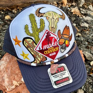 May include: A navy blue trucker hat with a white front panel featuring embroidered desert and cowboy-themed patches. The patches include a cactus, a bull skull, a cowboy skull wearing sunglasses, and the text "make America COWBOY AGAIN". A small charm of a cowboy hat hangs from the brim.