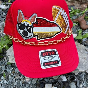 May include: Red trucker hat with a gold chain and patches. The patches include a cartoon dog wearing sunglasses, a football, and a sequined design. The hat has the text "OTTO WORLD FAMOUS FOAM FRONT TRUCKER".