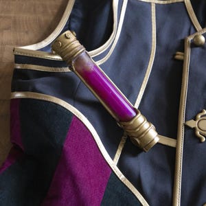 May include: A gold and black decorative vest with a gold trim and a small vial filled with a pink liquid.