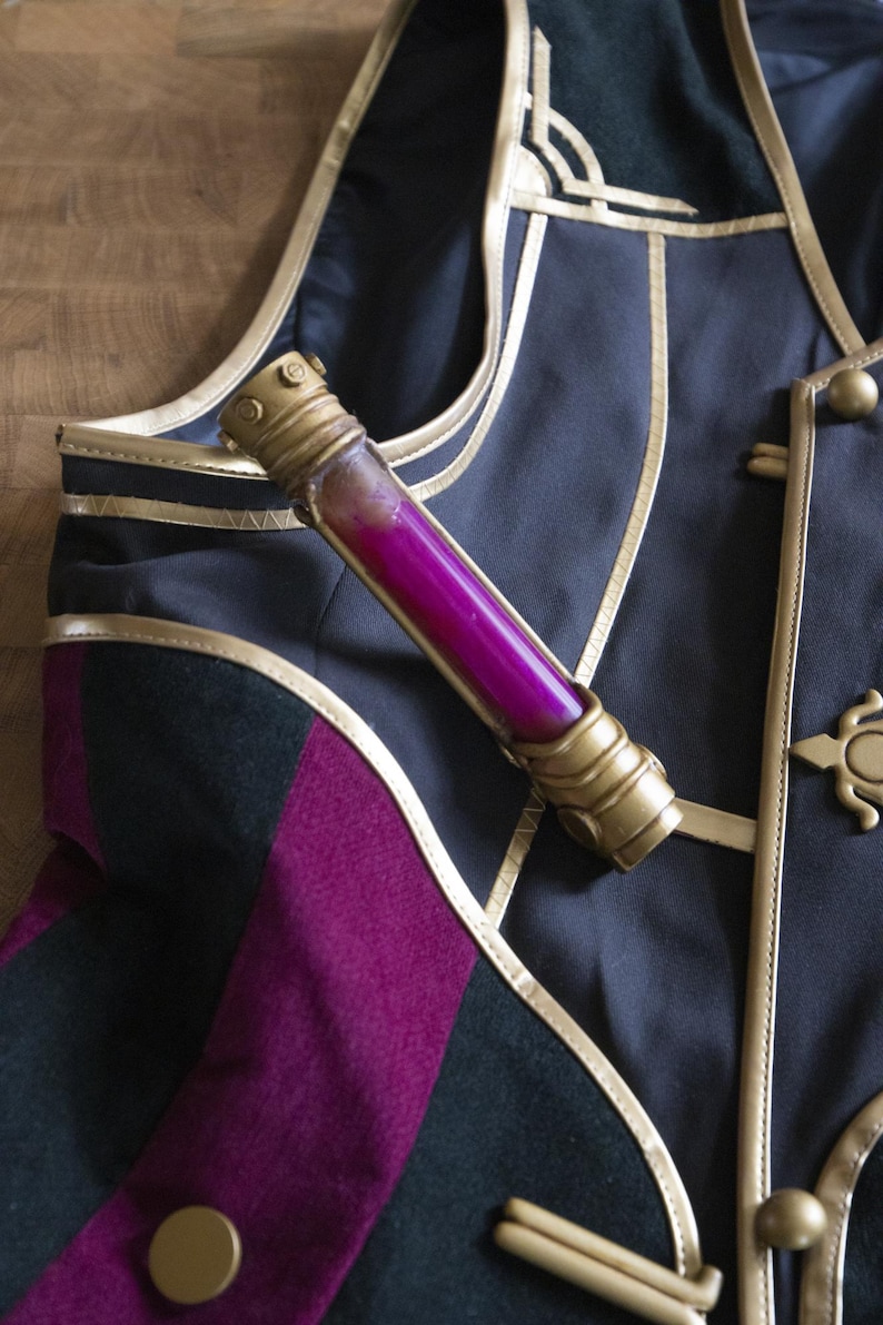 May include: A gold and black decorative vest with a gold trim and a small vial filled with a pink liquid.