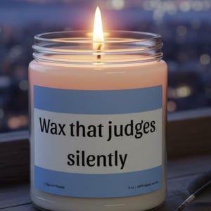 Puede incluir: Una vela encendida en un tarro de cristal con una etiqueta que dice "Wax that judges silently". La vela tiene cera de color melocotón y una llama brillante. Una pluma descansa sobre la superficie de madera junto a la vela. El fondo es una ciudad borrosa.